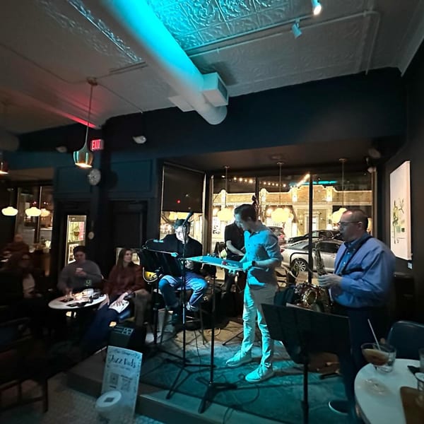A jazz band performs on a small stage in a dimly lit restaurant with patrons enjoying drinks.