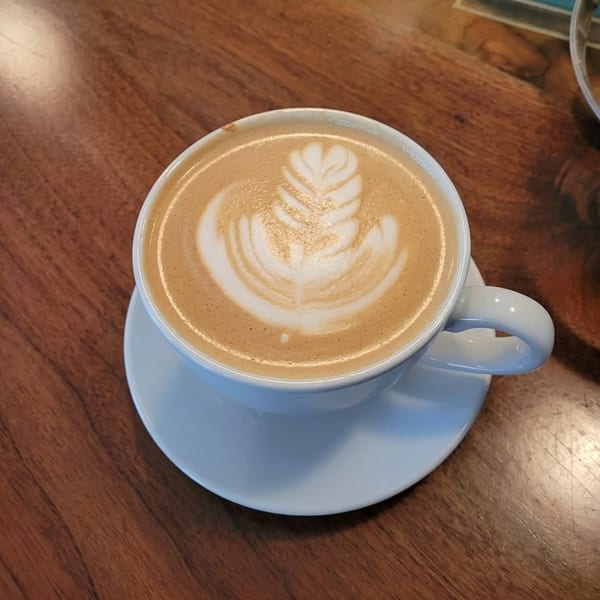 A white cup of latte with intricate leaf art in the foam, on a wooden table.