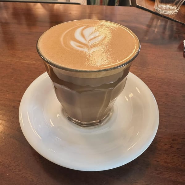 A latte with delicate latte art in a clear glass on a white saucer, on a wooden table.