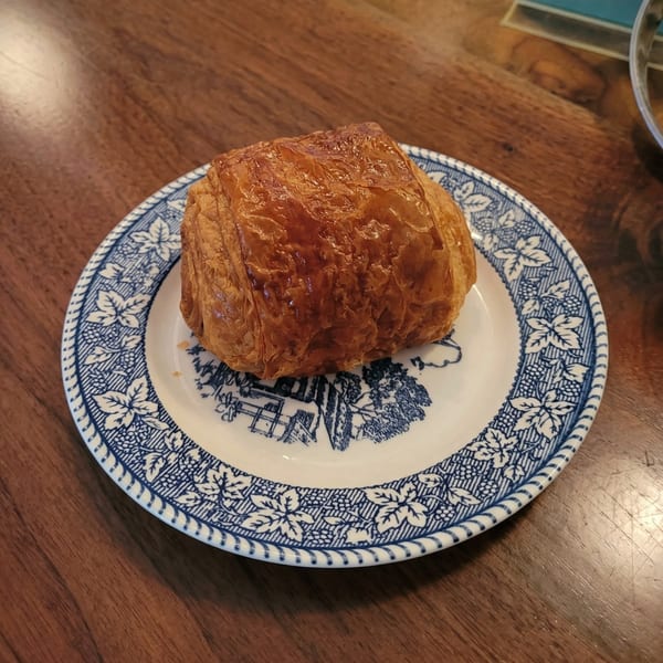 A golden-brown pain au chocolat sits on a white plate with blue floral patterns on a wooden table.