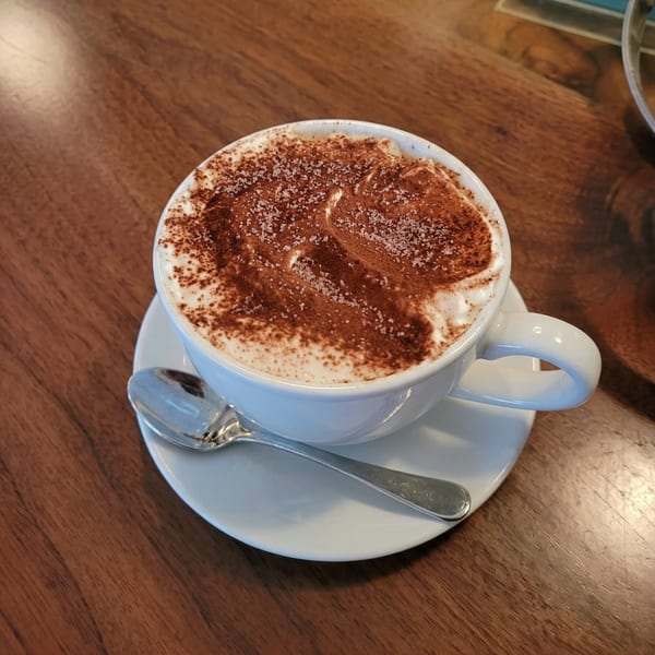 A white cup of cappuccino with whipped cream, cocoa powder, and sugar crystals on a wooden table.